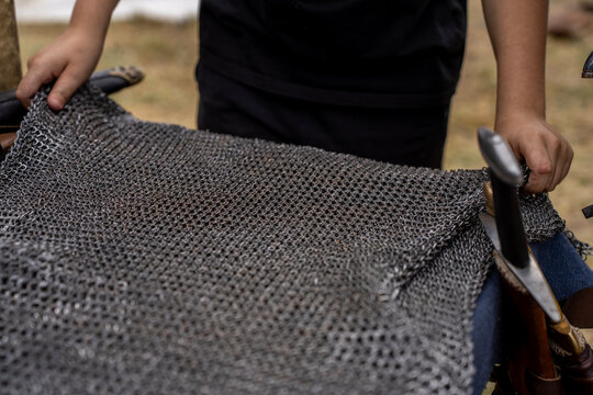 Close up of hands holding and displaying medieval chainmail armor fabric next to sheathed sword, showing steel ring mesh pattern and historical reenactment equipment.