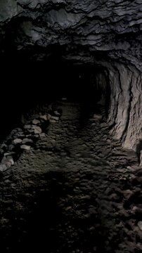 First-person point of view walking through a dark narrow cave tunnel with rough stone walls and dim light at the end.