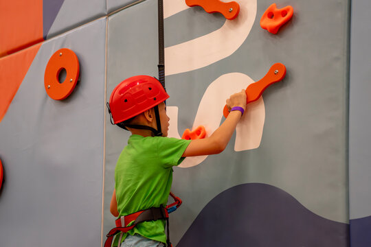 Boy in red safety helmet and harness climbing indoor bouldering wall with artificial grips, practicing rock climbing sport and active recreation in entertainment center.