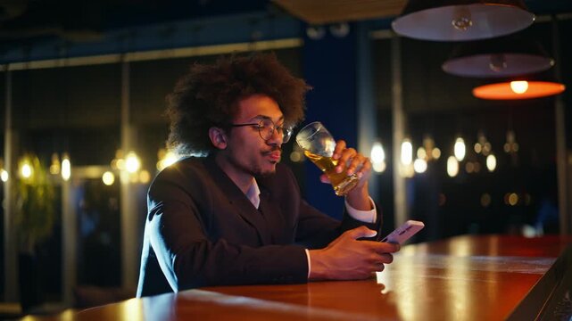 Employee sipping beer bar counter checking smartphone. Man smiling night pub.