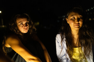 Two women sitting together at night, engaged in conversation under dim lighting near a waterfront © KONSTANTIN SHISHKIN