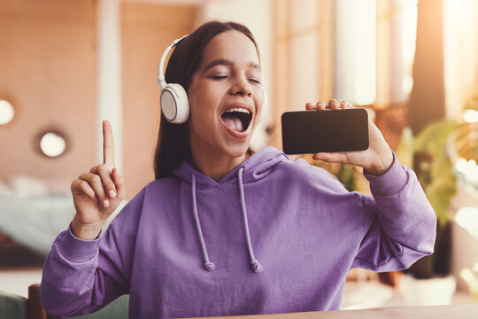 Happy Young Latina Female Enjoying Music While Singing Along With Smartphone, Sitting Indoors In Cozy Home Environment. Cheerful Millennial Woman, Engaging In Fun And Relaxation.