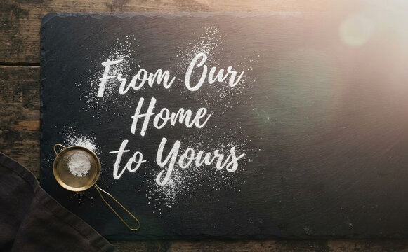 From Our Home to Yours text written with white flour on a dark slate board with a sifter and cloth background concept for baking and cooking recipe background