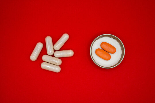 White pills and capsules spilling from open medicine bottle. Pharmaceutical tablets and supplements for health treatment and wellness on red backgr...