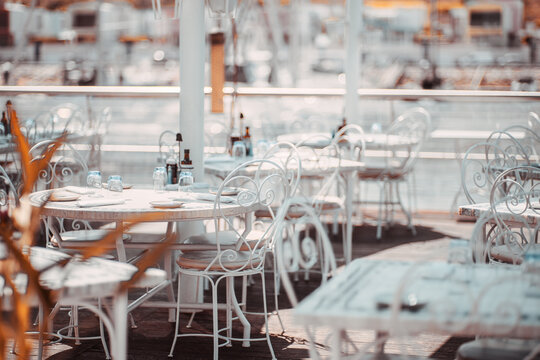 Selective focus on an elegant empty waterfront dining terrace with white wrought iron chairs, set tables, sparkling glassware and softly blurred marina in warm morning light