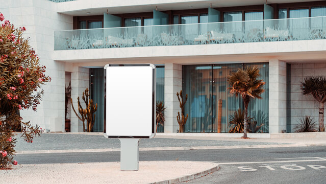 Blank vertical advertising template on a freestanding roadside display before a modern resort facade with glass balconies, tropical landscaping and clear copy space