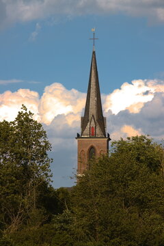 Historic brick church tower steeple rising behind green trees under dramatic evening sky