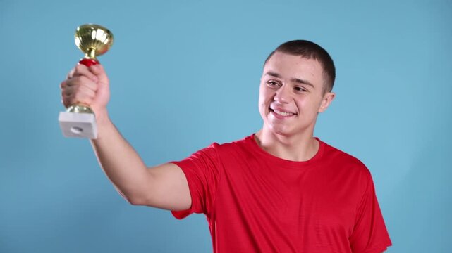 Happy teenage boy holding gold trophy cup against blue background. Camera moving, slow motion