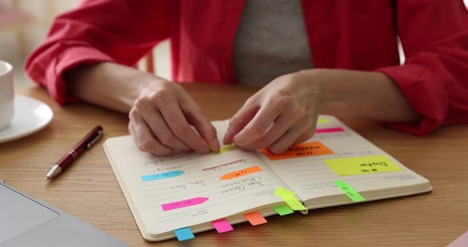 Planning. Woman putting stickers in notebook at wooden desk indoors, closeup