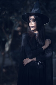 Halloween theme: A young woman with ghostly makeup and hair, dressed as a witch, holding a magic wand and a pumpkin amidst a graveyard; Halloween festival concept.