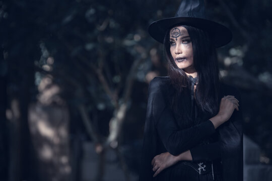 Halloween theme: A young woman with ghostly makeup and hair, dressed as a witch, holding a magic wand and a pumpkin amidst a graveyard; Halloween festival concept.
