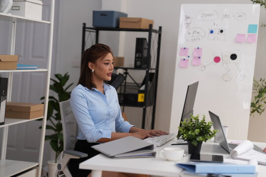 Reacting to work on screen with Asian businesswoman at desk with monitor and papers; later in a modern office, medium shot, real people at work, everyday business life
