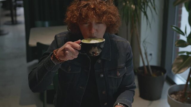 Person with curly hair sips matcha drink in a cafe, seated at a table with plants and a relaxed atmosphere enhancing the experience