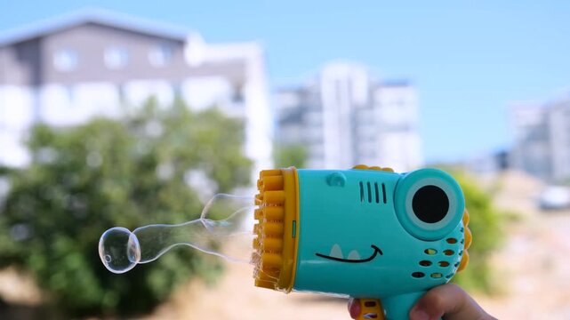 Slow motion side angle video of an automatic blue and yellow plastic bubble blower gun firing transparent soap bubbles outdoors in bright sunny daylight.