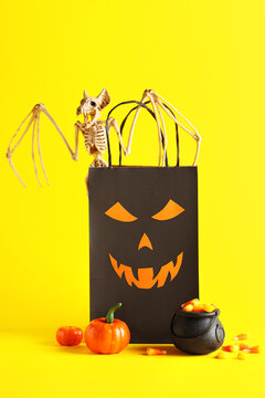 Cauldron with tasty Halloween candy corns, pumpkin, bat skeleton and paper bag on yellow background