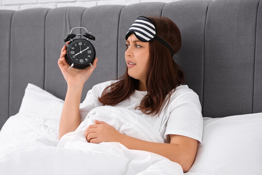 Annoyed young woman with alarm clock lying in bed at morning