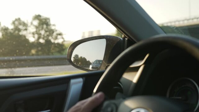 Driver's hand turns the steering wheel while adjusting the side mirror view of a passing vehicle on a bright highway surrounded by greenery