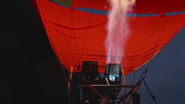 Hot air balloon burner releases flames, lighting up the red canopy as it prepares for takeoff against a darkening sky