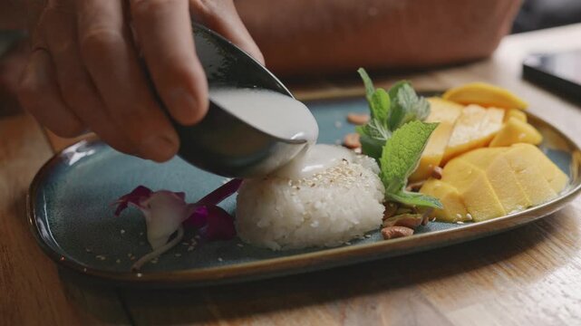 Hand drizzles coconut milk onto sticky rice dessert with mango slices and mint leaves on a blue plate, creating a visually appealing dish