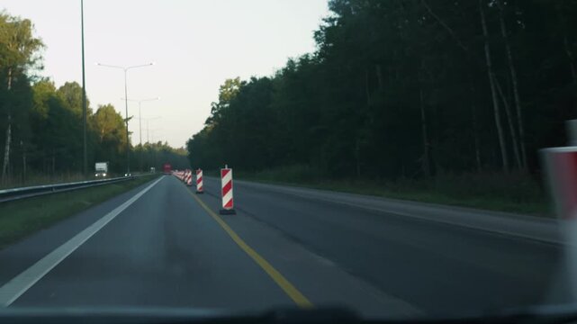 Traffic cones are placed along a highway, with vehicles passing by and trees visible, illustrating the ongoing road construction process