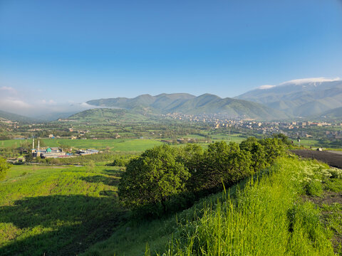 Rolling green hills lead toward snow-dusted peaks, a khanqah rising in the near valley with a city stretching farther beyond, framed by a lush hedge of trees and tall grass.
📍 MahmoudAbad, Sarvabad