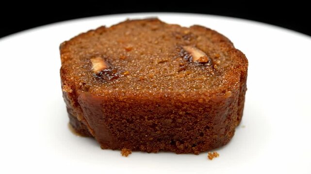 Delicious close-up of a moist date cake or sticky toffee pudding slice, rich brown dessert texture with nuts and glaze, served on a bright white plate.