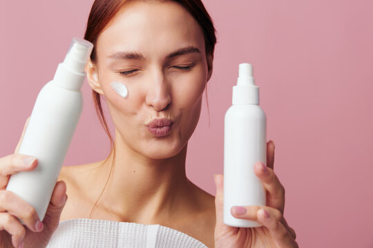 Playful young woman with puckered lips holds two white cosmetic spray bottles toward viewer, cream swatch on cheek. Studio shot on pink background. Fun beauty and skincare promotion.