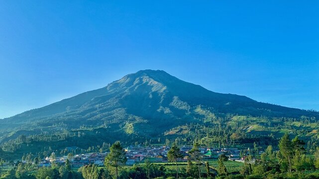 Landscape view of Mount Sumbing with countryside village settlement surrounded by green hills under clear blue sky in Central Java, Indonesia.