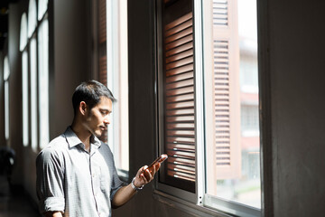 Fototapeta premium Man standing by bright corridor windows using a smartphone, illuminated by natural light and shadows, wearing a casual shirt, focused on the screen inside a modern building.