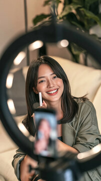 Beauty Influencer Recording Tutorial: A young woman smiles radiantly while holding a makeup product, filming a beauty tutorial with a ring light and smartphone, creating engaging content online.