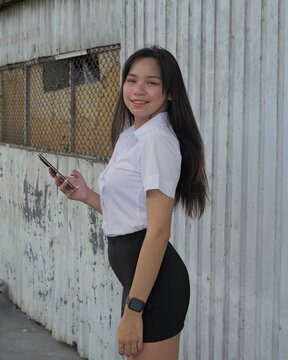 A young Asian woman in a student uniform holds a smartphone by a corrugated metal wall