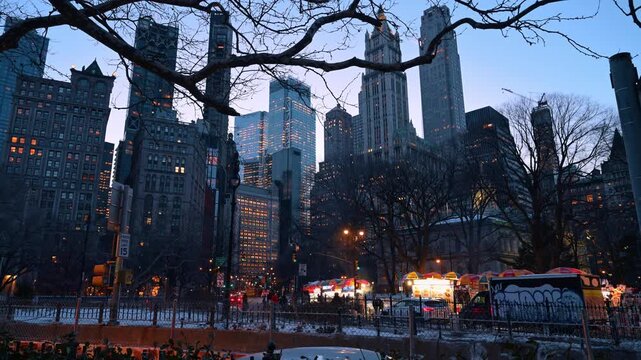 Lower Manhattan skyline and street food carts at dusk. Lit windows rise behind bare branches, food carts and a parked truck beside patches of snow.