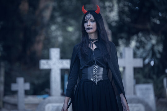 Halloween theme: A young woman with ghostly makeup and hair, dressed as a witch, holding a magic wand and a pumpkin amidst a graveyard; Halloween festival concept.