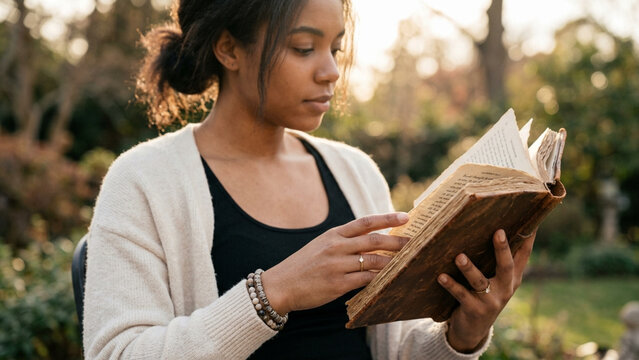 A young thoughtful woman deeply concentrated while reading a captivating novel outdoors under warm afternoon sunlight surrounded by natural greenery during a peaceful golden hour moment