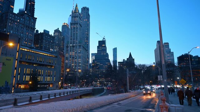 New York, USA, 9 January 2026 : Pedestrians walking along winter path near Lower Manhattan skyscrapers. People move on snowy sidewalk beside lit street lamps as evening sky turns blue.