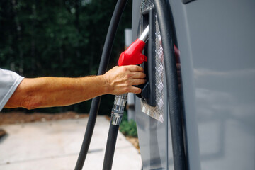 Hand with a red fuel nozzle near a silver car during refueling at an open gas station. The black hose and the reflective side panel are visible. Fuel concept. © Кирилл Горшков