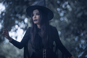 Halloween theme: A young woman with ghostly makeup and hair, dressed as a witch, holding a magic wand and a pumpkin amidst a graveyard  Halloween festival concept. © ThawKyar