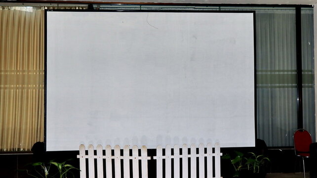 Large blank projector screen surrounded by a white fence, dark seating, curtains, and greenery in a formal indoor meeting venue prepared for presentations. now.