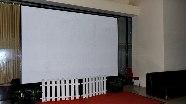 Blank projection screen in a conference room with a white picket fence, dark sofas, curtains, and plants forming a simple indoor venue backdrop for formal uses.