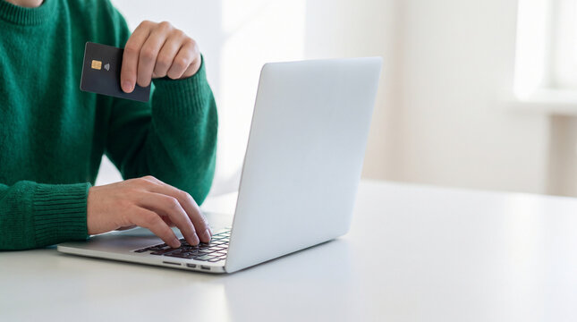 Man in green sweater holding credit card and typing on laptop for online shopping, close up of hands making secure digital payment transaction on white table, e-commerce concept