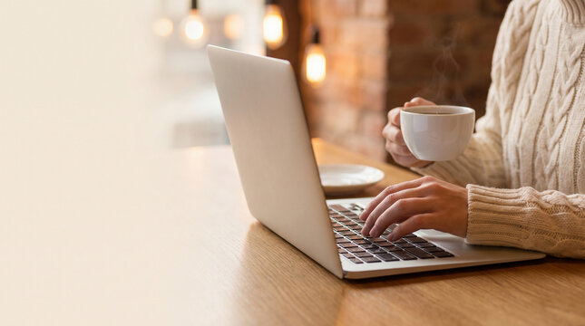 Woman in cozy sweater holding hot coffee cup while working on laptop, close up of female hands typing on keyboard at wooden table in cafe, remote freelance work lifestyle concept