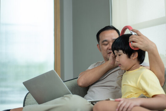 Affectionate Asian father putting red wireless headphones on his little boy for online learning or entertainment on laptop. Family lifestyle concept. Human-created, not AI.