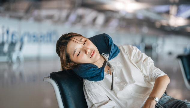 Tired asian woman passenger wearing a travel pillow takes a nap on a chair in the airport terminal waiting flight delay departure