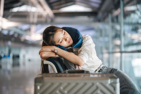 Tired asian woman passenger wearing a travel pillow takes a nap on a chair in the airport terminal waiting flight delay departure