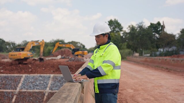 Asian male civil engineer using laptop computer and radio transceiver to supervise bridge construction. Heavy civil infrastructure engineering, technology and site management concept.