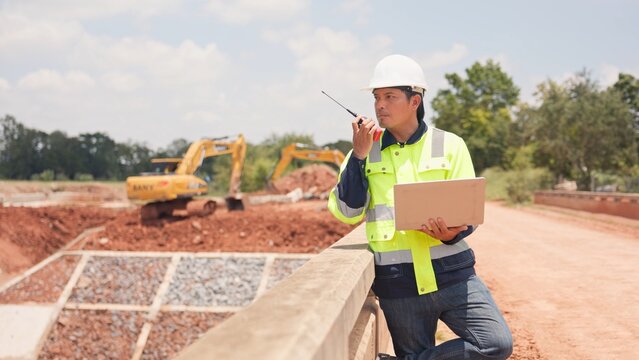 Asian male civil engineer using laptop computer and radio transceiver to supervise bridge construction. Heavy civil infrastructure engineering, technology and site management concept.
