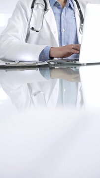Unrecognizable doctor man in a blue shirt under a medical coat is using technology in medicine. Close-up of hands typing on a laptop on a glass desk in clinic, vertical view. Health care theme concept