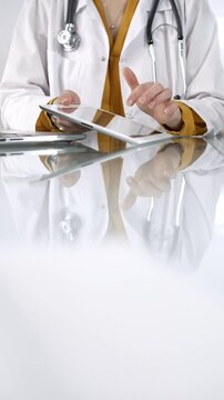 Unknown doctor woman wearing a stethoscope, yellow blouse under a lab coat, is using digital tablet in modern healthcare setting, interacting with patient data, vertical portrait. Medicine concept
