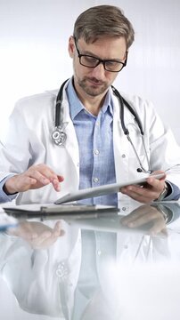Doctor man in blue shirt under a white coat, glasses and stethoscope is using digital tablet in modern healthcare setting, interacting with patient data, vertical portrait view. Medicine and science