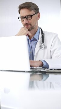Doctor man in a blue shirt under a medical coat is thinking while using technology in medicine. Portrait of physician typing on a laptop on a glass desk in clinic, vertical view. Medicine and science